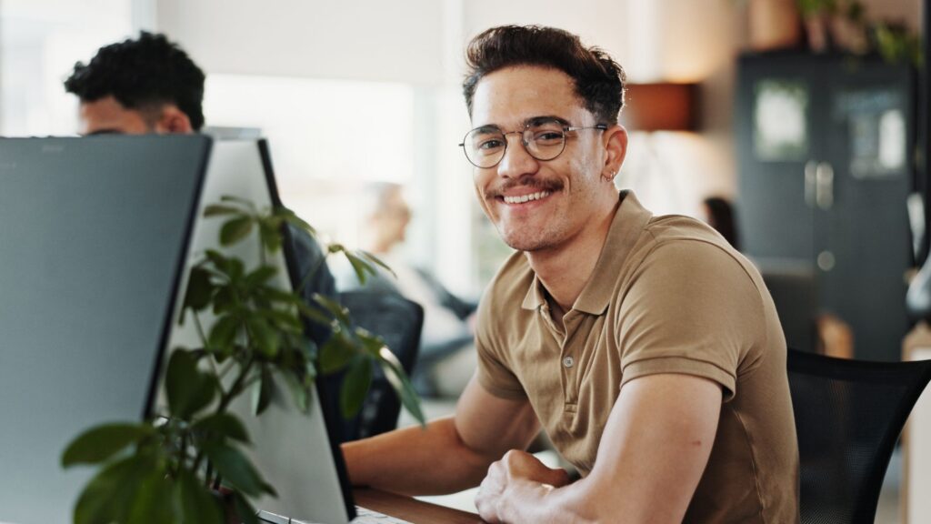 Lächelnder Mitarbeiter sitzt im Büro am Computerarbeitsplatz und blickt in die Kamera; im Hintergrund sind weitere Kolleginnen und Kollegen zu sehen.